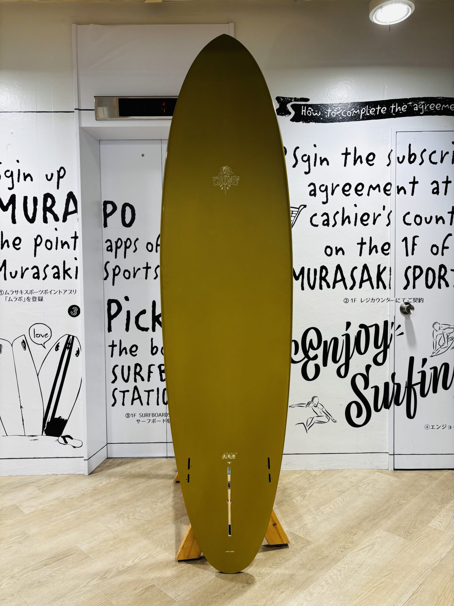 Mustard-yellow surfboard upright on a wooden rack indoors, with bold wall art in the background.
