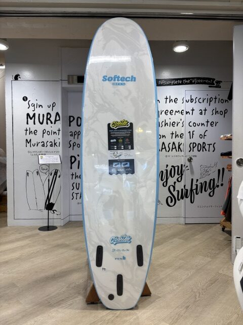 White surfboard with blue edge on a display rack in a shop, Softech logo near the top and a product label in the center.