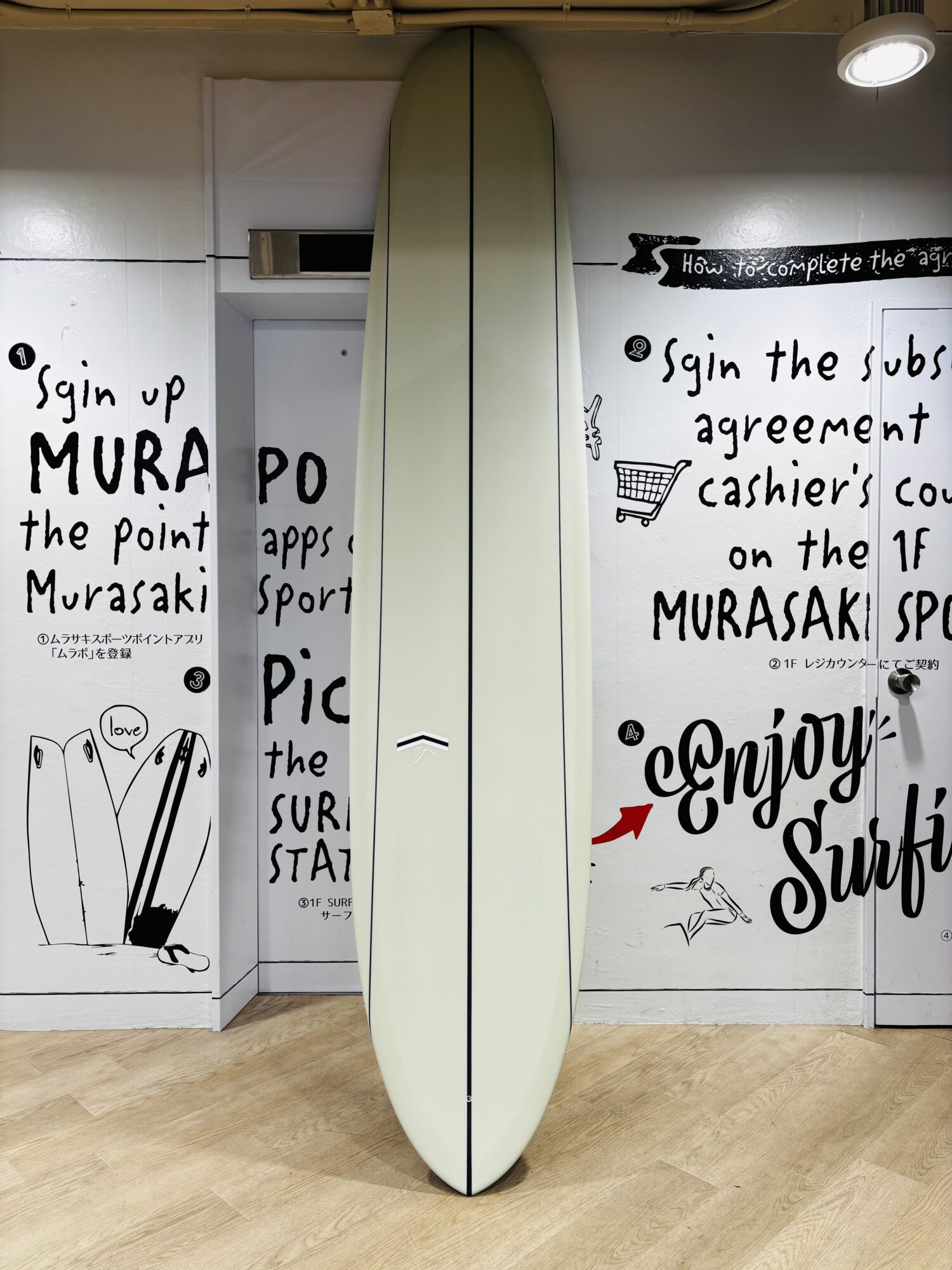 Cream-colored surfboard standing upright between white wall panels with bold black handwritten text graphics and small illustrations on the walls.