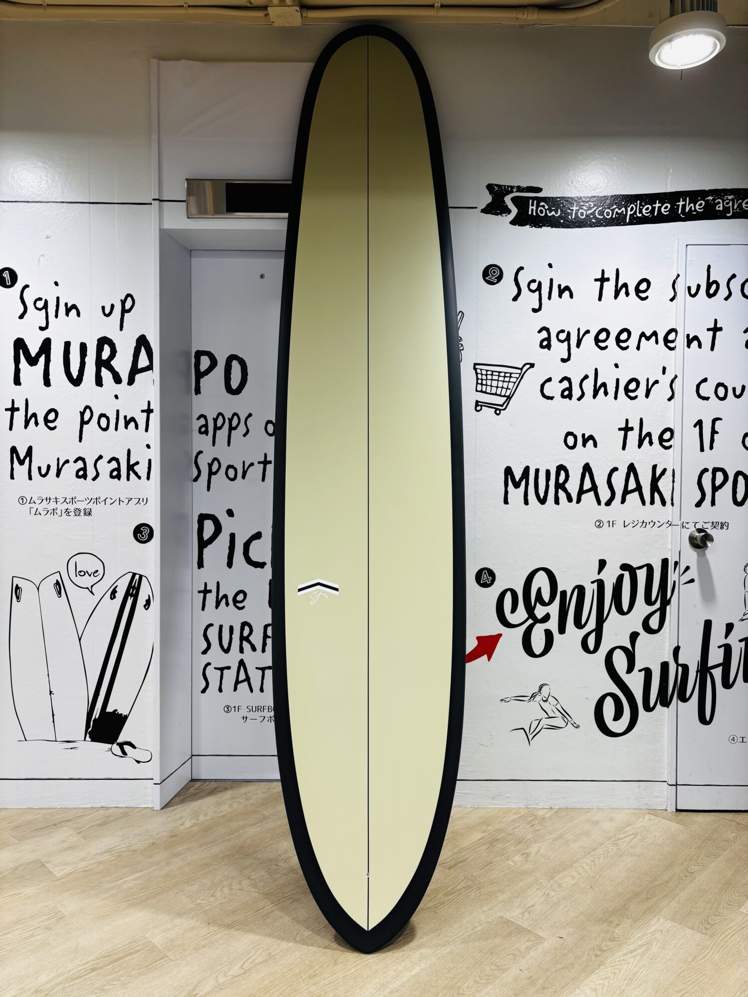 Tall yellow surfboard with a black edge standing upright indoors against walls covered in black handwritten text and drawings on a white backdrop.