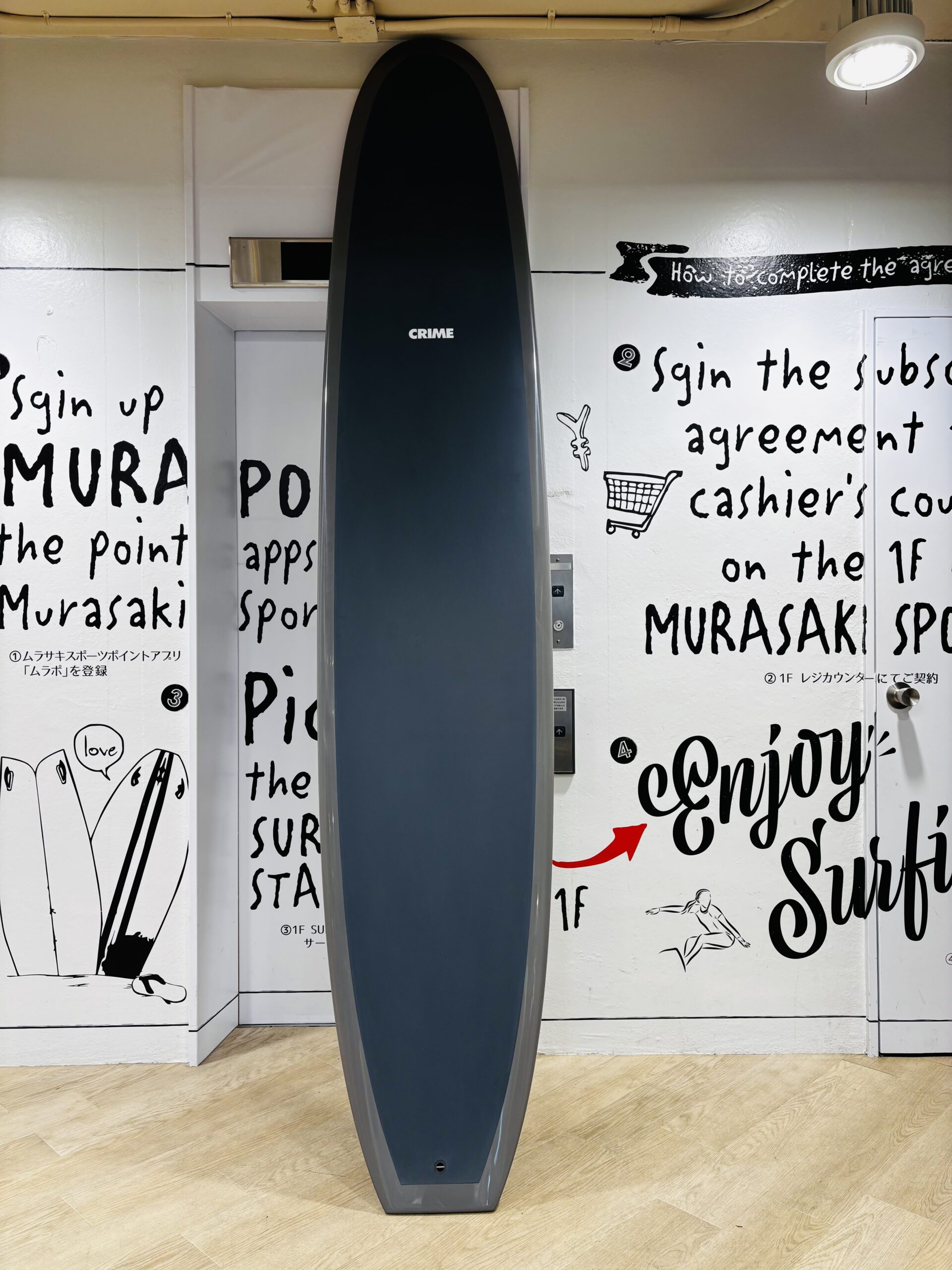 Tall dark blue surfboard standing upright between white walls covered with black handwritten text and drawings in a gallery-like setting.