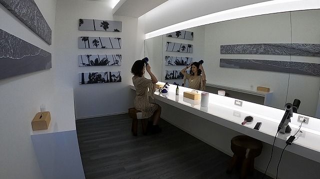 bdc8507dccbd348af7a577dbb10a417f - SHOP NEWS | ムラサキスポーツオンラインストア 通販 Woman at a bright white makeup vanity applying makeup while looking in the large mirror; wall shelves display black-and-white palm tree photos.