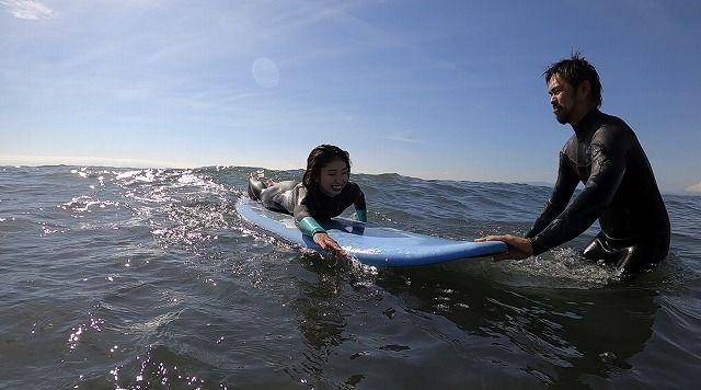 da91a83e465784033a8b79cf8062d672 - SHOP NEWS | ムラサキスポーツオンラインストア 通販 A young girl lies on a blue surfboard riding a small wave while a man in a wetsuit supports the board in the water next to her.