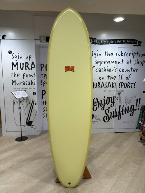 Yellow longboard surfboard mounted on a wooden rack inside a surf shop, with surf-themed posters on the walls.