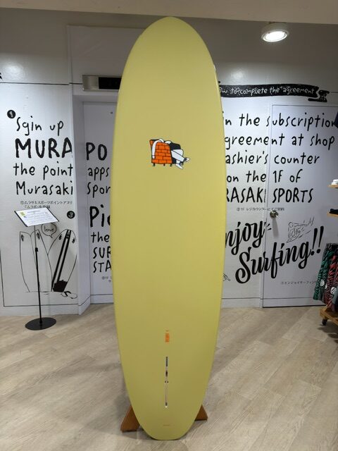 Tall pale-yellow surfboard on display in a shop, mounted upright on a wooden stand with a small cartoon patch near the center edge of the board.