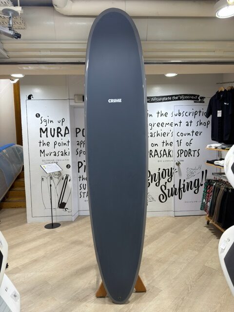 Gray stand-up surfboard standing upright on a wooden rack inside a shop, with the word 'CRIME' on its surface and merchandise visible in the background.