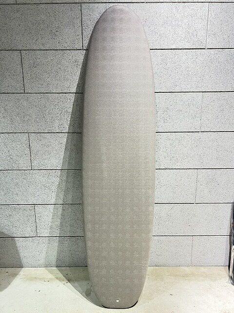 Light gray surfboard with rounded nose standing upright against a gray brick wall area.
