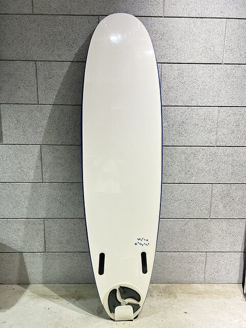 White surfboard with a rounded nose standing upright against a gray brick wall; two small fin slots near the tail and a single fin box visible at the bottom area