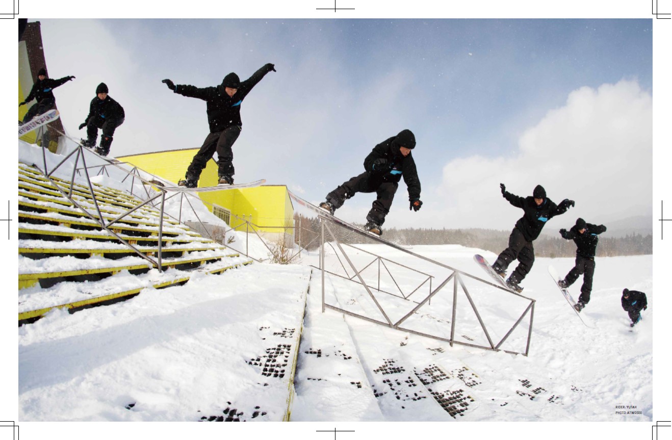Several snowboarders in black jackets glide along a metal rail over a snowy course, with a yellow ramp in the background and a clear sky.