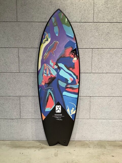 Colorful abstract surfboard leaning against a gray tiled wall, featuring blue, purple, red shapes and a black grip at the bottom.