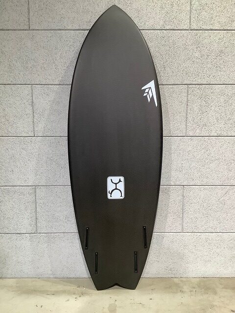 Black surfboard standing upright against a gray tiled wall, featuring a white logo near the upper half and four fin boxes at the bottom.