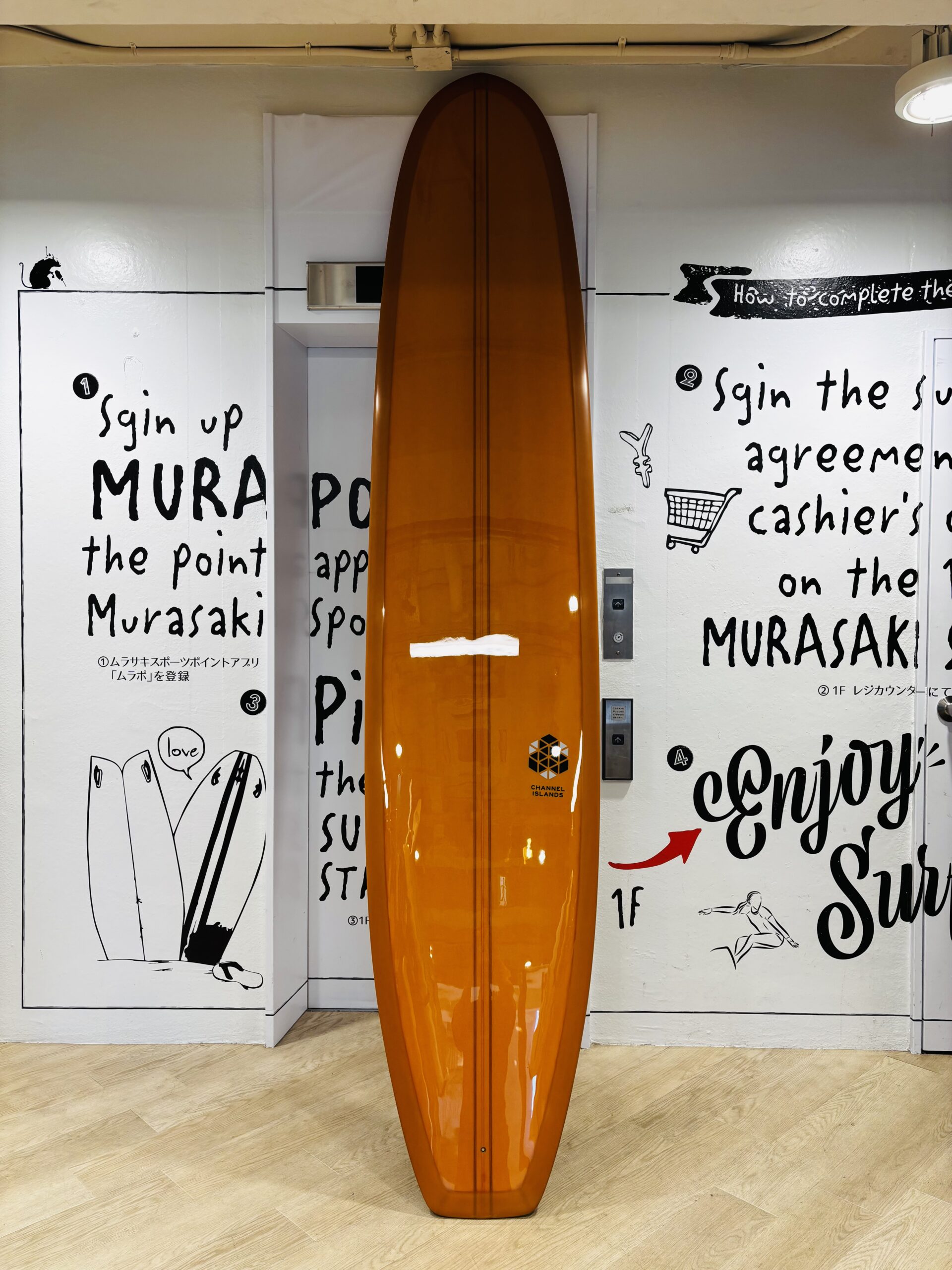 Tall orange surfboard standing upright in a white-walled room with black graphic text and drawings on the walls behind it.