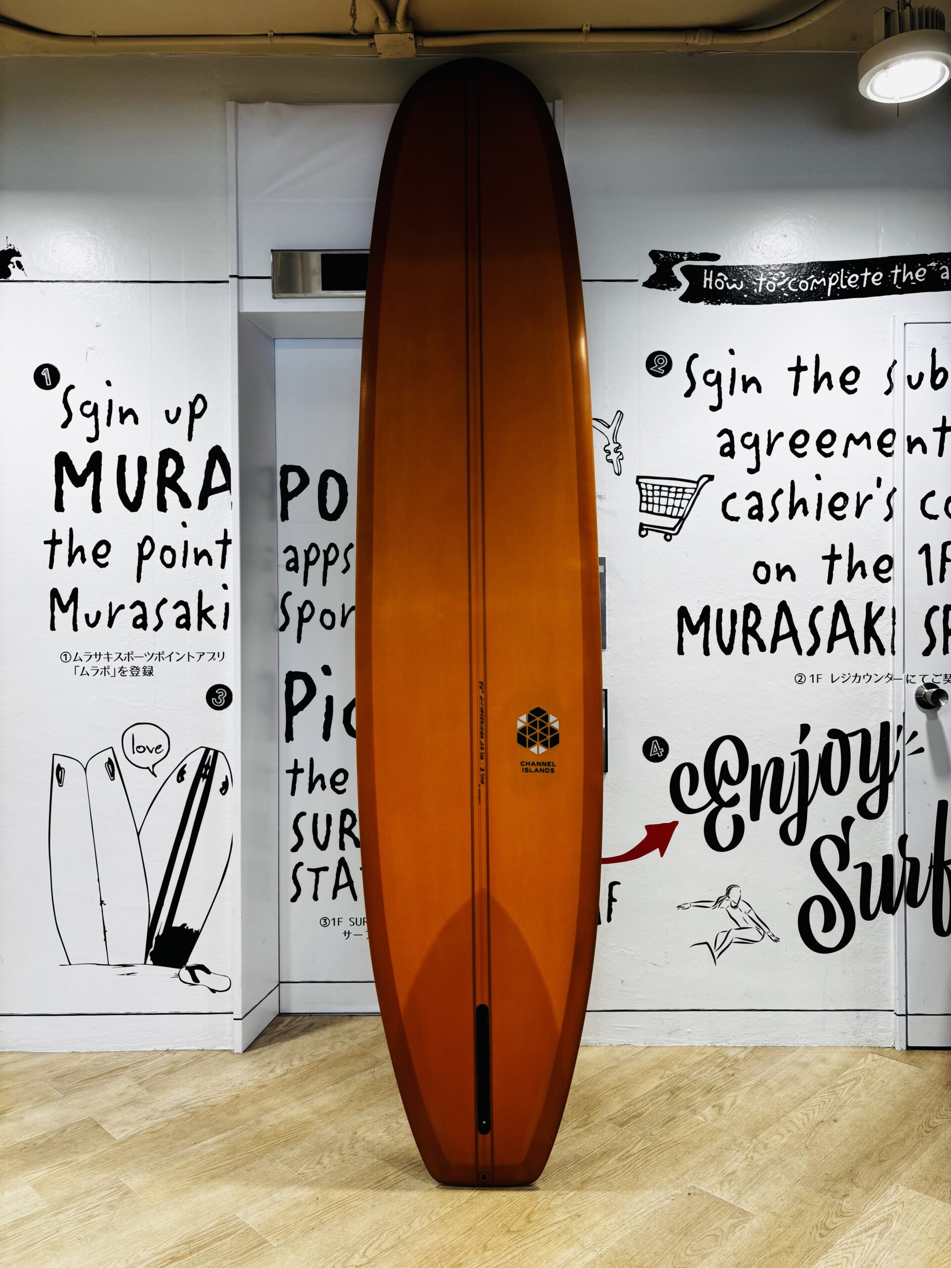 Orange surfboard standing upright indoors against a white wall covered in black handwritten text and drawings, with a wooden floor beneath.