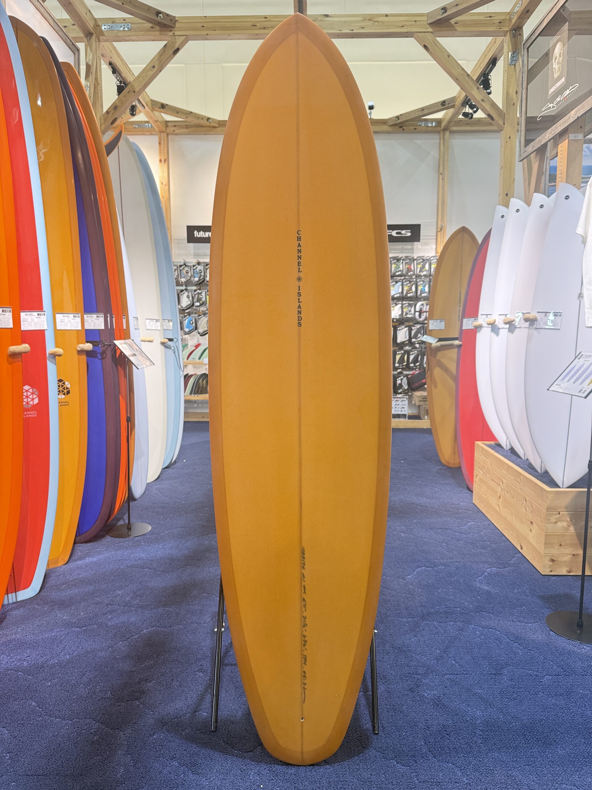 Orange surfboard standing upright on a display stand, with colorful boards lining both sides in a surf shop.