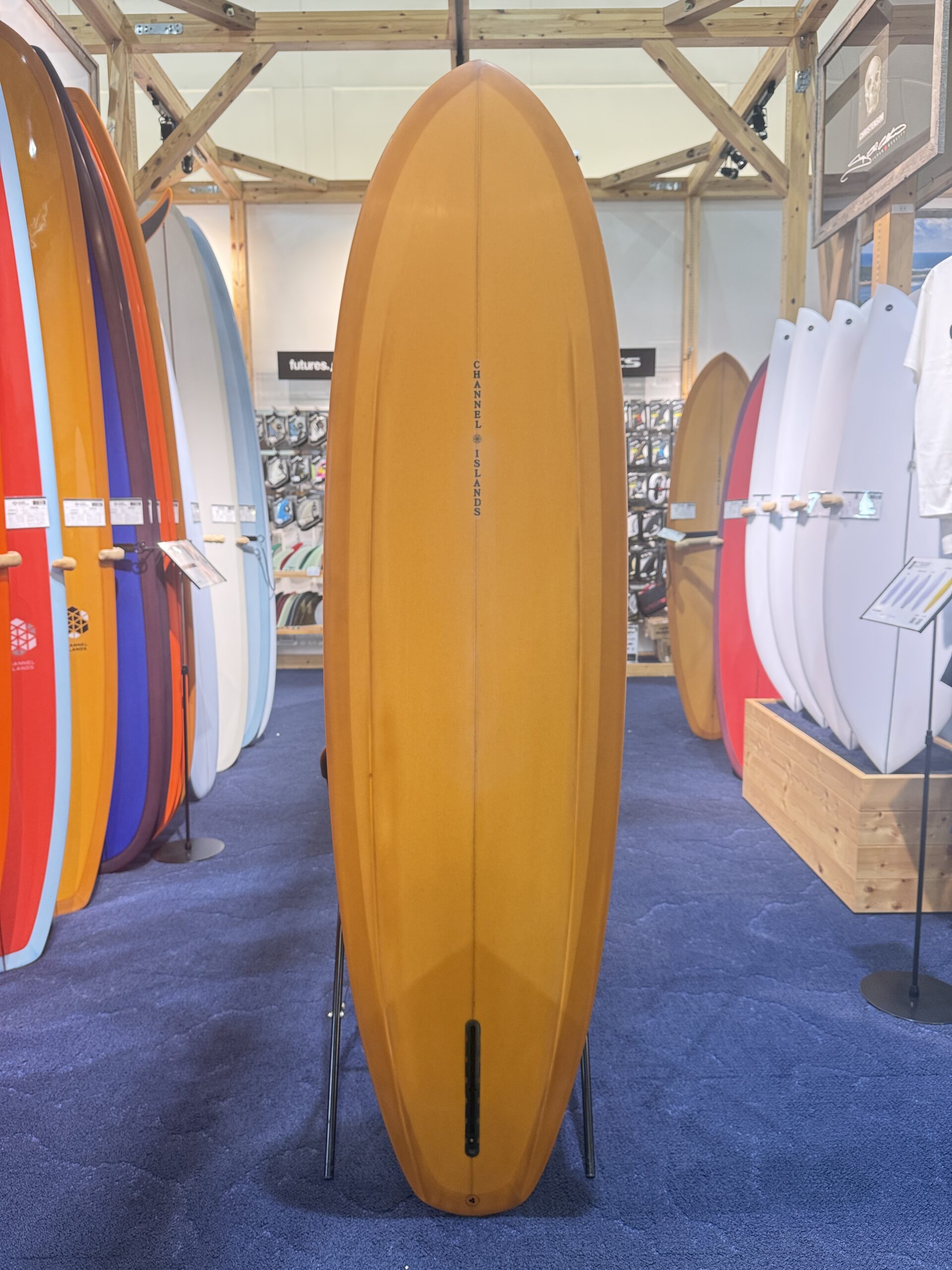 Tall orange surfboard on a stand in a surf shop, surrounded by colorful boards on racks.