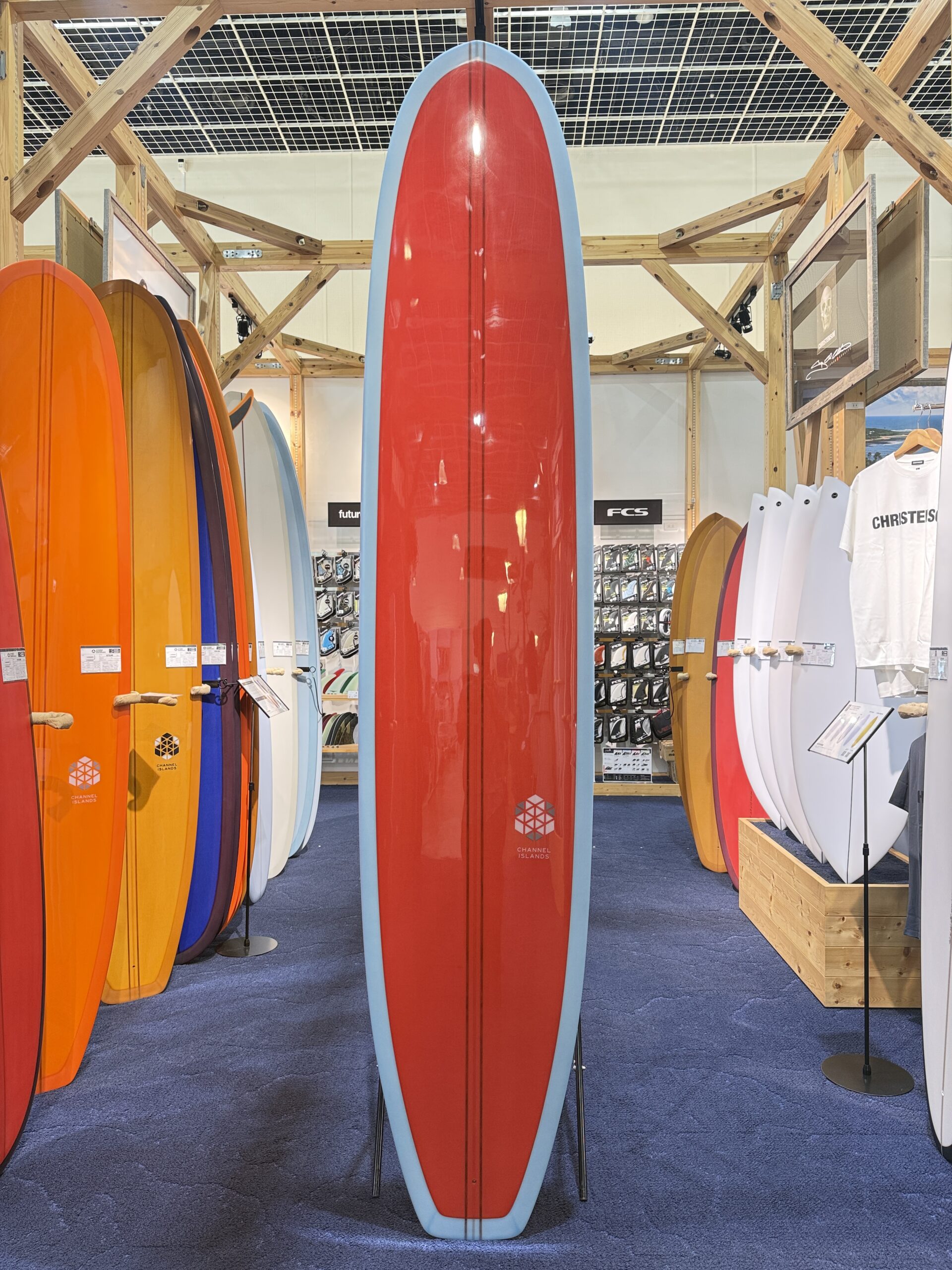 IMG_3920 - SHOP NEWS | ムラサキスポーツオンラインストア 通販 Tall red surfboard with light blue edge standing upright in a store display among orange, yellow, and white boards.