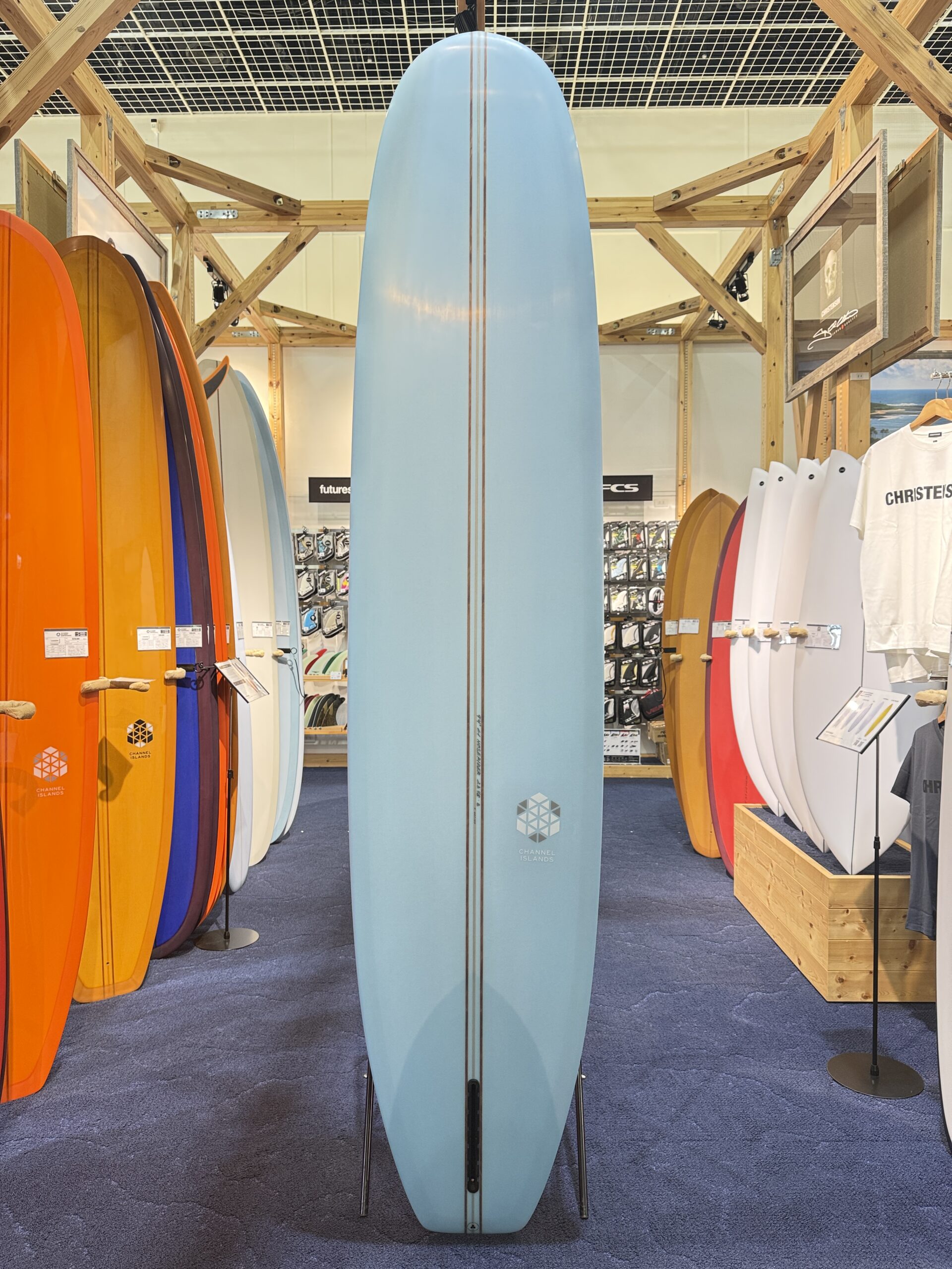 IMG_3921 - SHOP NEWS | ムラサキスポーツオンラインストア 通販 Center blue longboard with brown pinstripes stands upright on metal legs in a surf shop, surrounded by colorful boards and wooden display racks, with the Channel Islands logo toward the base.