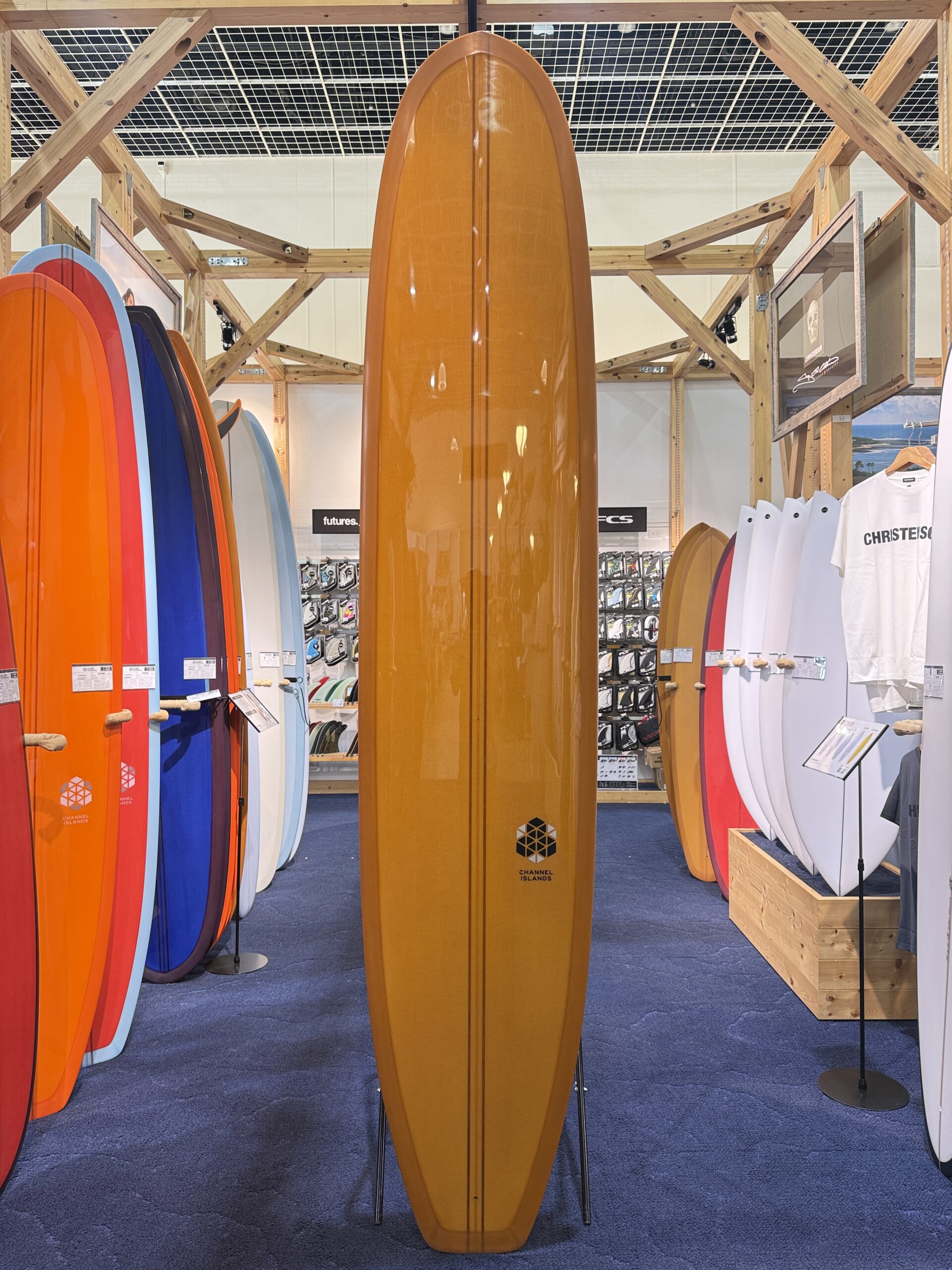IMG_3922 - SHOP NEWS | ムラサキスポーツオンラインストア 通販 Tall orange surfboard upright in a shop aisle, with colorful boards lined along both sides.