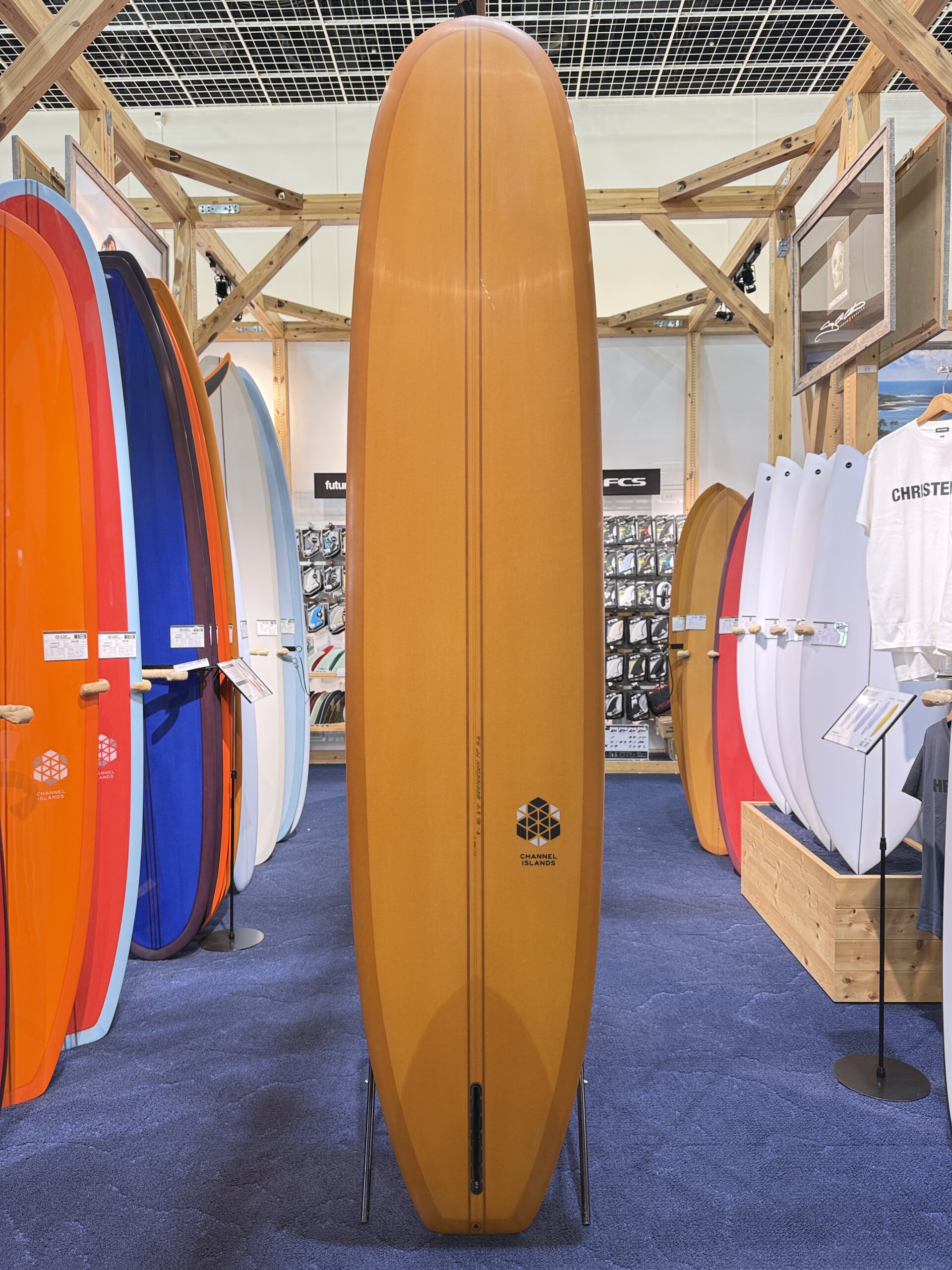 IMG_3924 - SHOP NEWS | ムラサキスポーツオンラインストア 通販 Tall orange surfboard standing upright in a surf shop display, surrounded by colorful boards on racks and a wooden frame structure behind it.