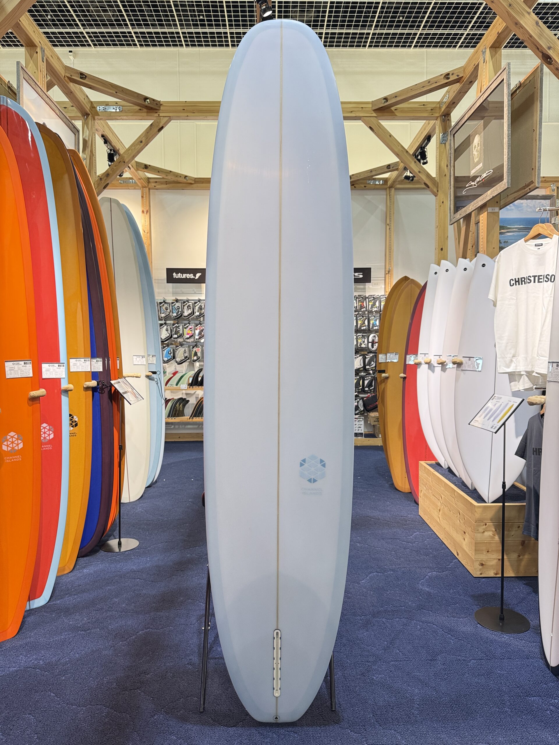 Tall pale-blue longboard standing upright on a display stand in a surf shop, with colorful boards lined up on both sides behind it.