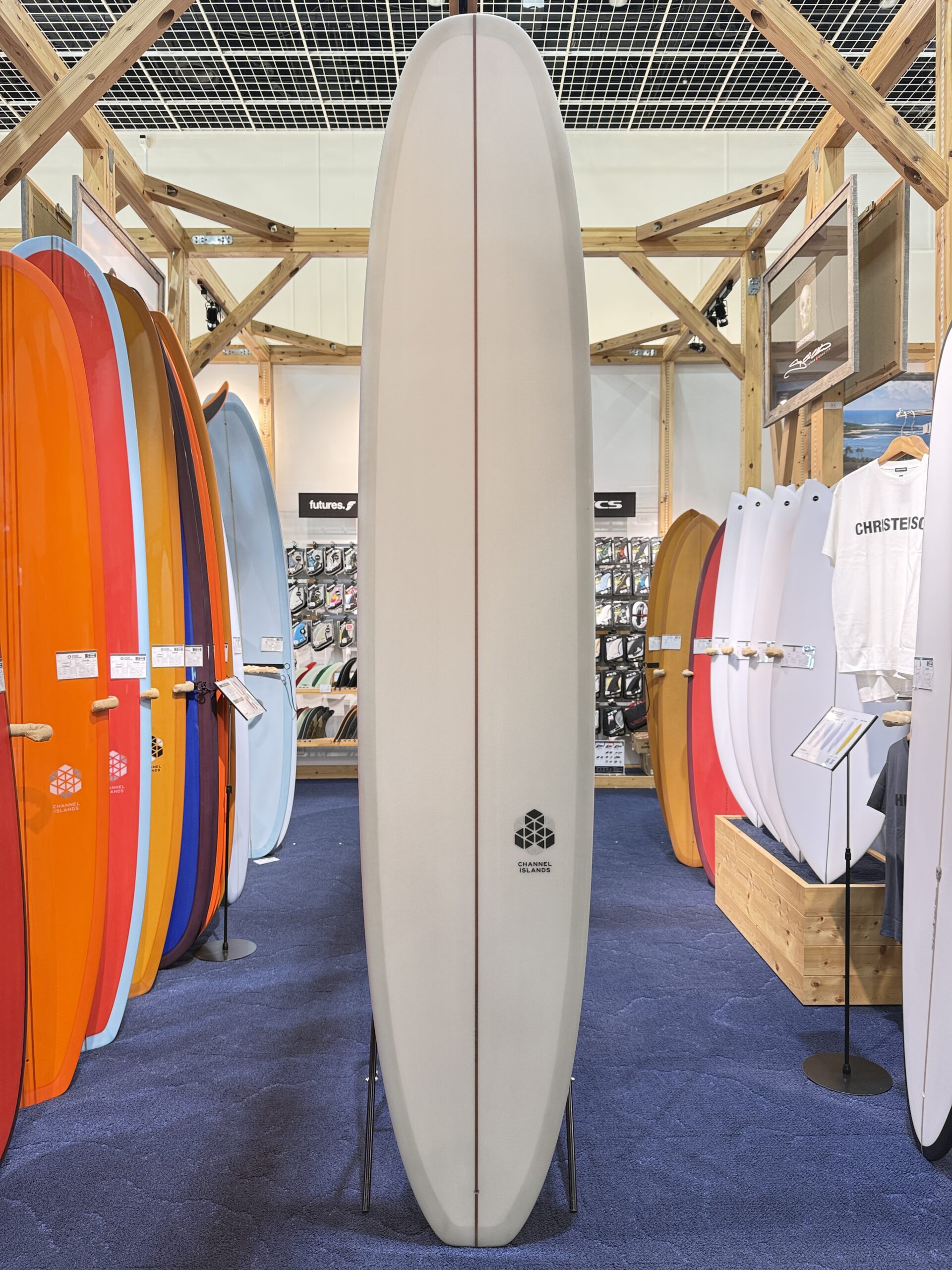 White surfboard standing upright in a shop with colorful boards on both sides and wooden display racks around it.