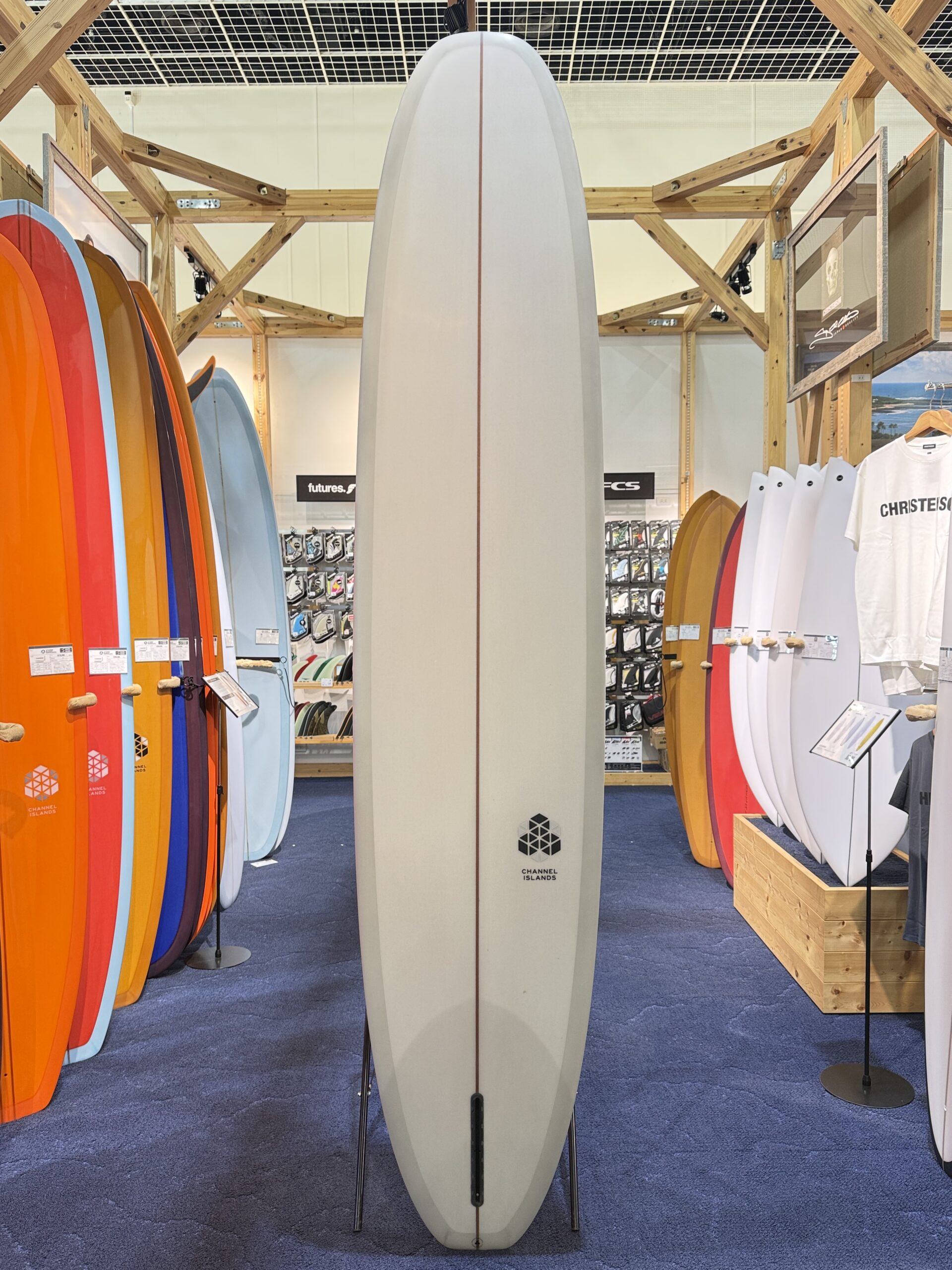 Tall white surfboard with a brown stringer standing upright on a display stand, centered in a surf shop with colorful boards on either side.