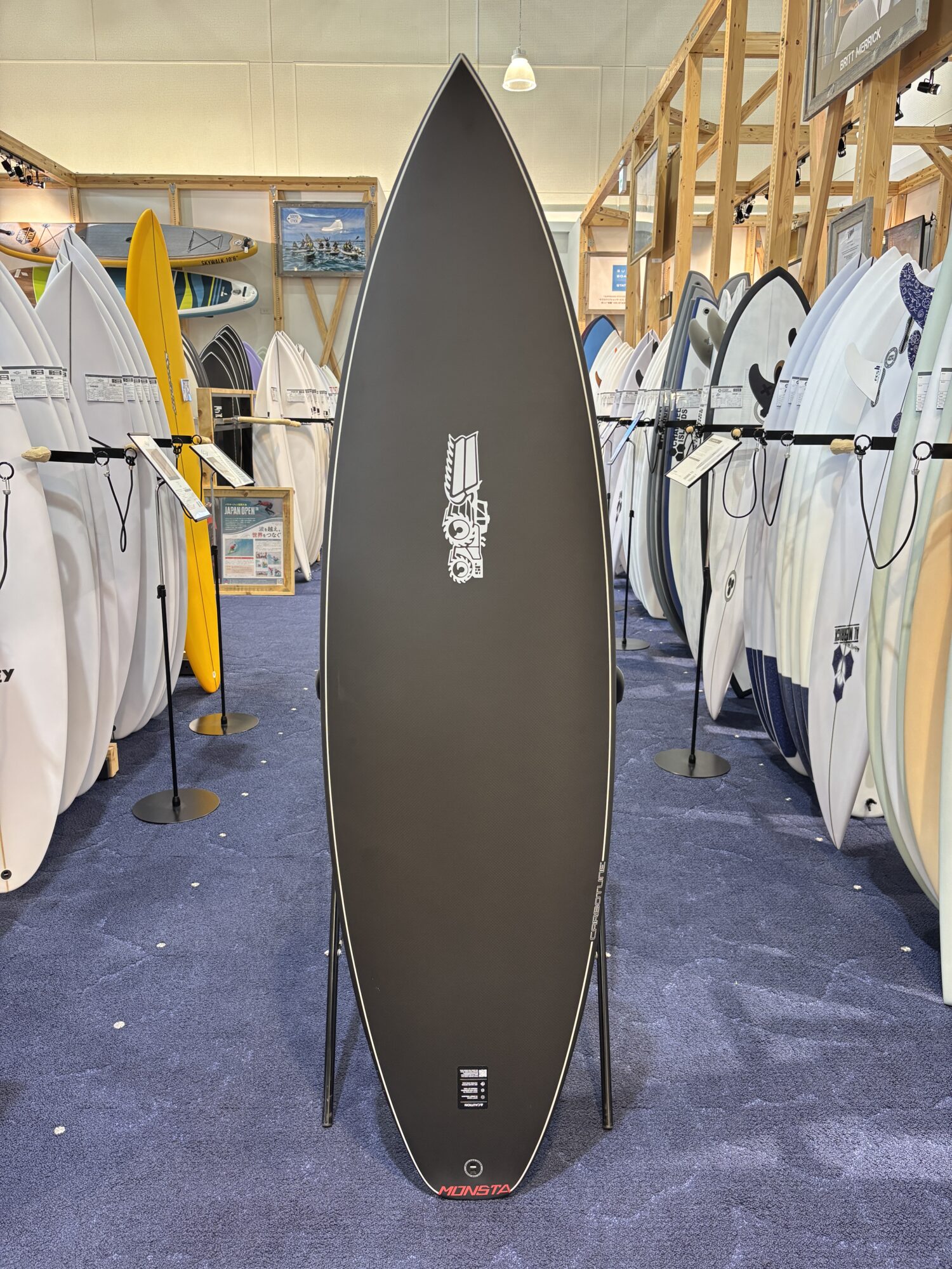 Tall black surfboard displayed upright in a showroom, surrounded by white and colored boards on wooden racks.