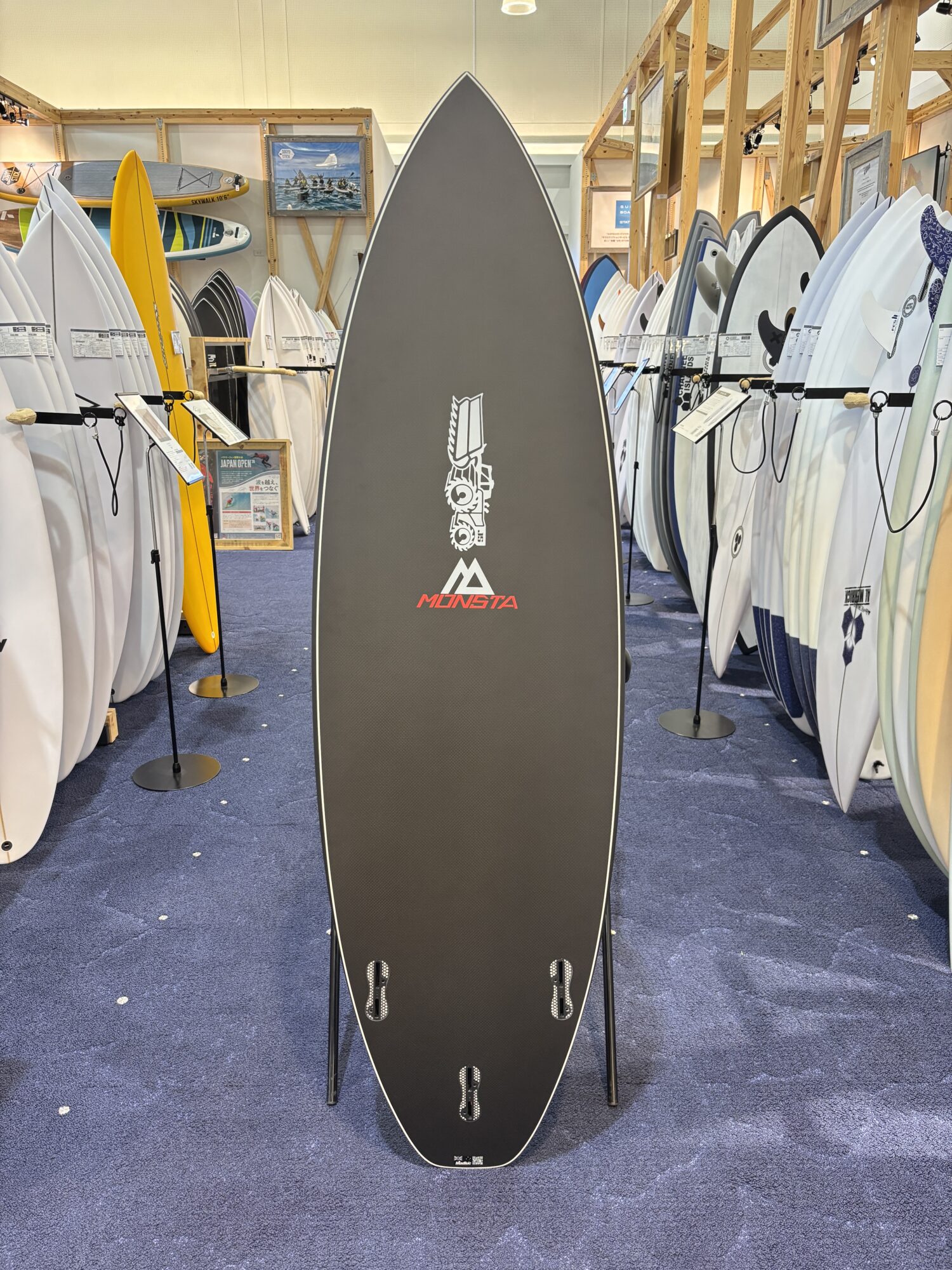 Indoor surf shop with rows of boards on racks; a tall dark Monsta longboard centered in the foreground.