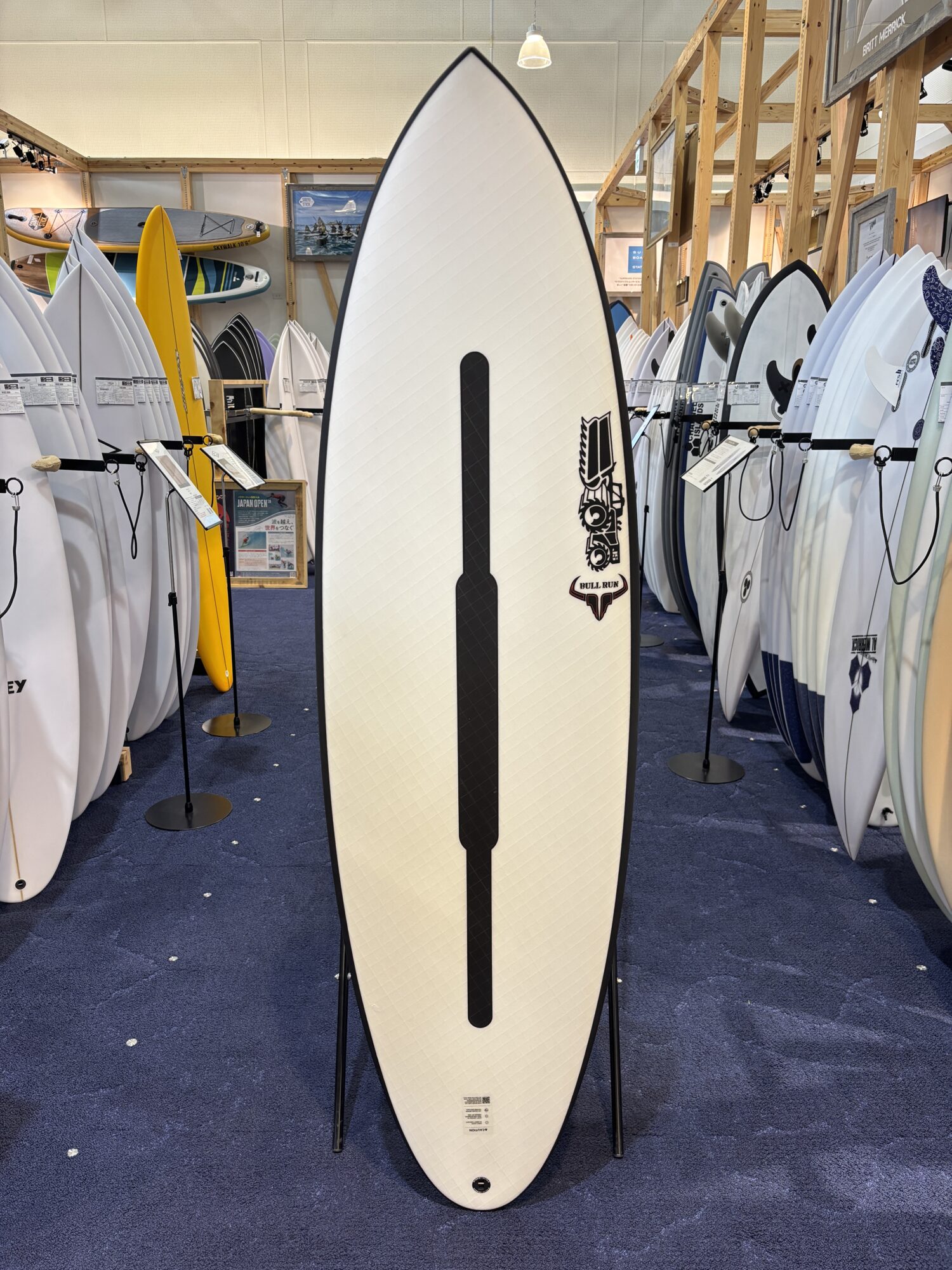 Tall white surfboard with a black vertical stripe on display in a showroom full of boards in a shop.