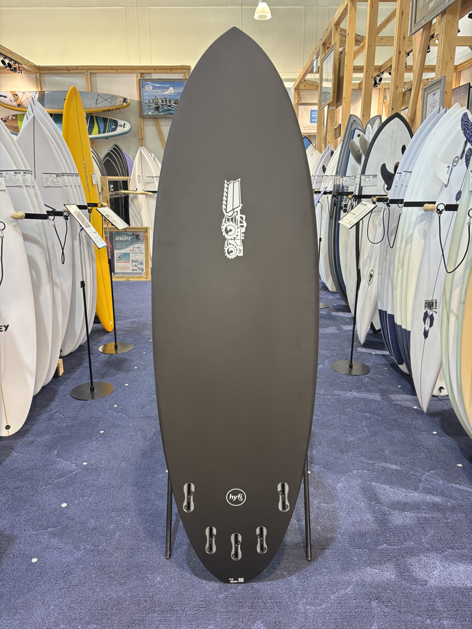 Tall dark gray surfboard standing upright in a showroom, surrounded by racks of white and colored boards and a yellow board in the background.