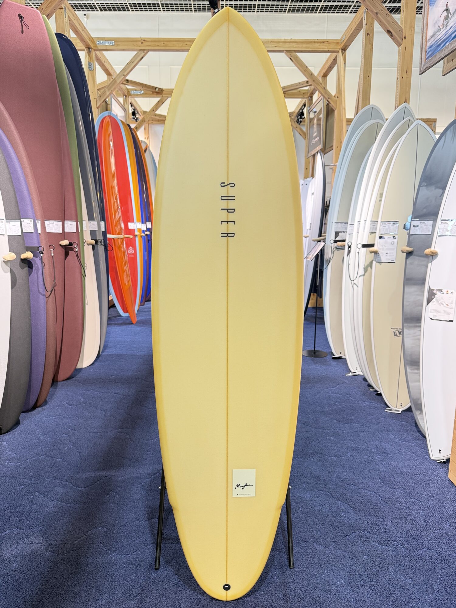 IMG_4017 - SHOP NEWS | ムラサキスポーツオンラインストア 通販 Yellow longboard surfboard on a display stand in a showroom, surrounded by colorful boards on racks.