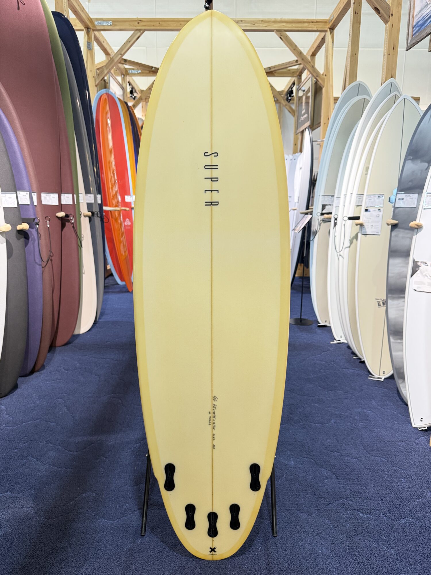 IMG_4018 - SHOP NEWS | ムラサキスポーツオンラインストア 通販 Yellow surfboard with four fins on a stand in a surf shop, flanked by colorful boards on racks.