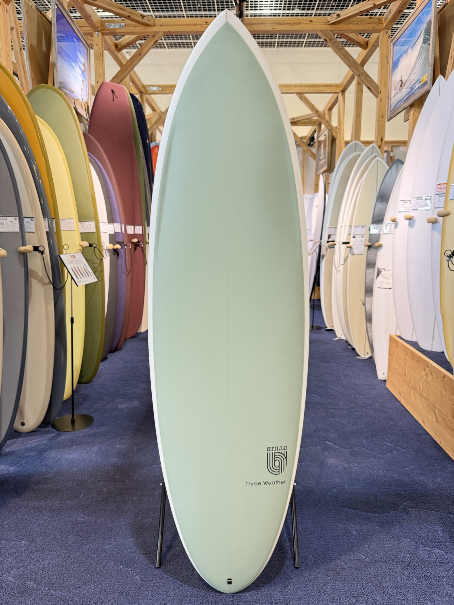 IMG_4021 - SHOP NEWS | ムラサキスポーツオンラインストア 通販 Pastel green surfboard on a display stand in a showroom, with a row of colorful boards lined along wooden racks on both sides.