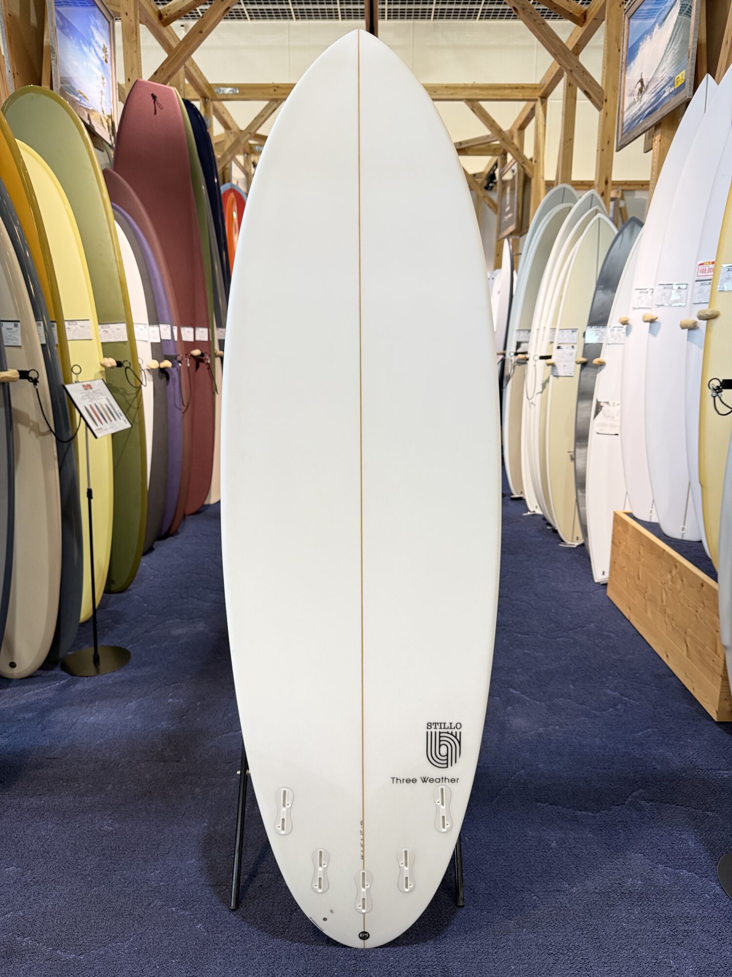 IMG_4022 - SHOP NEWS | ムラサキスポーツオンラインストア 通販 Center-front white surfboard with a brown stringer, displayed upright in a store surrounded by colorful boards.