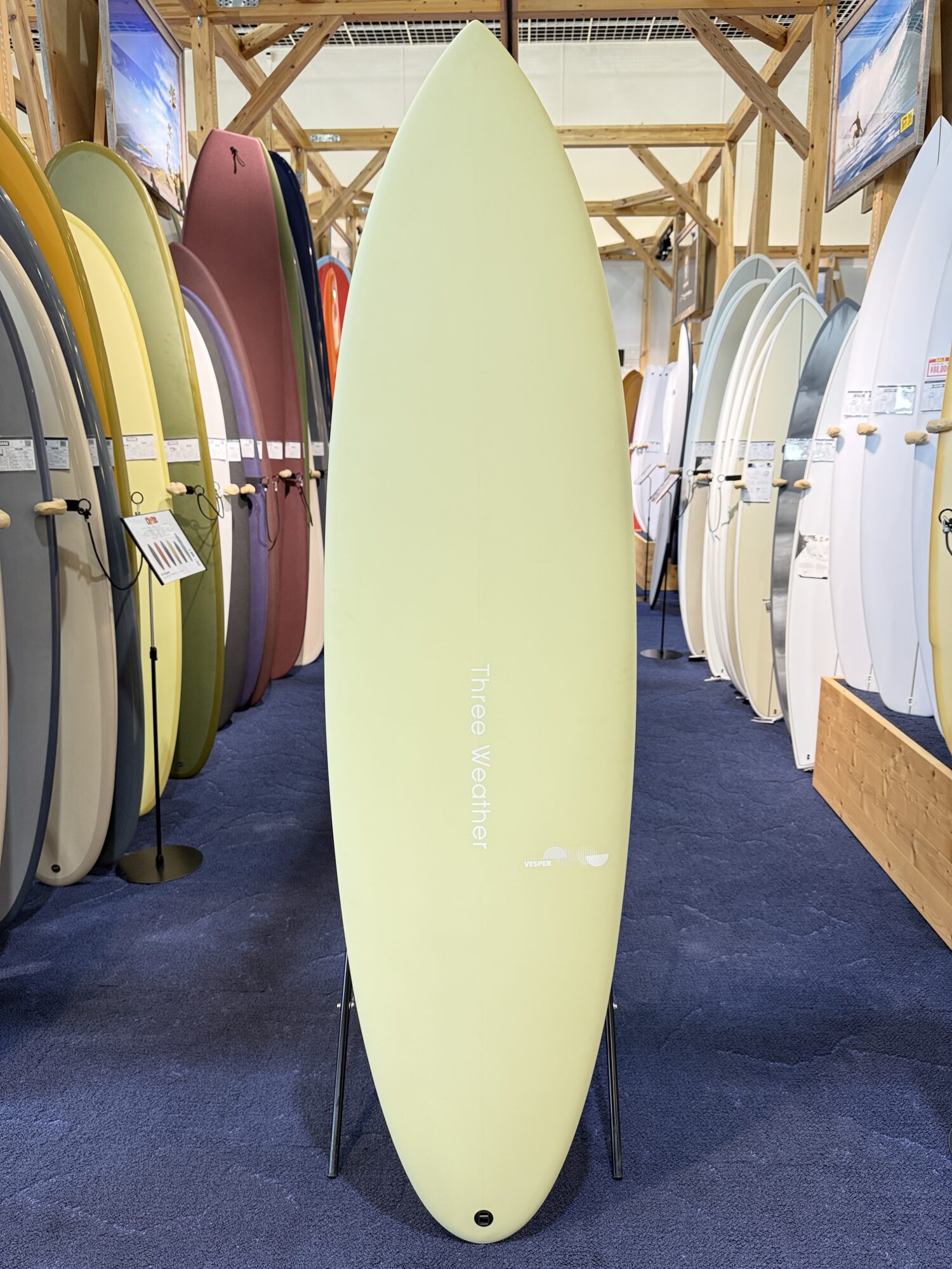 IMG_4023 - SHOP NEWS | ムラサキスポーツオンラインストア 通販 Pale green surfboard on a stand in a showroom, surrounded by many colorful longboards on both sides.