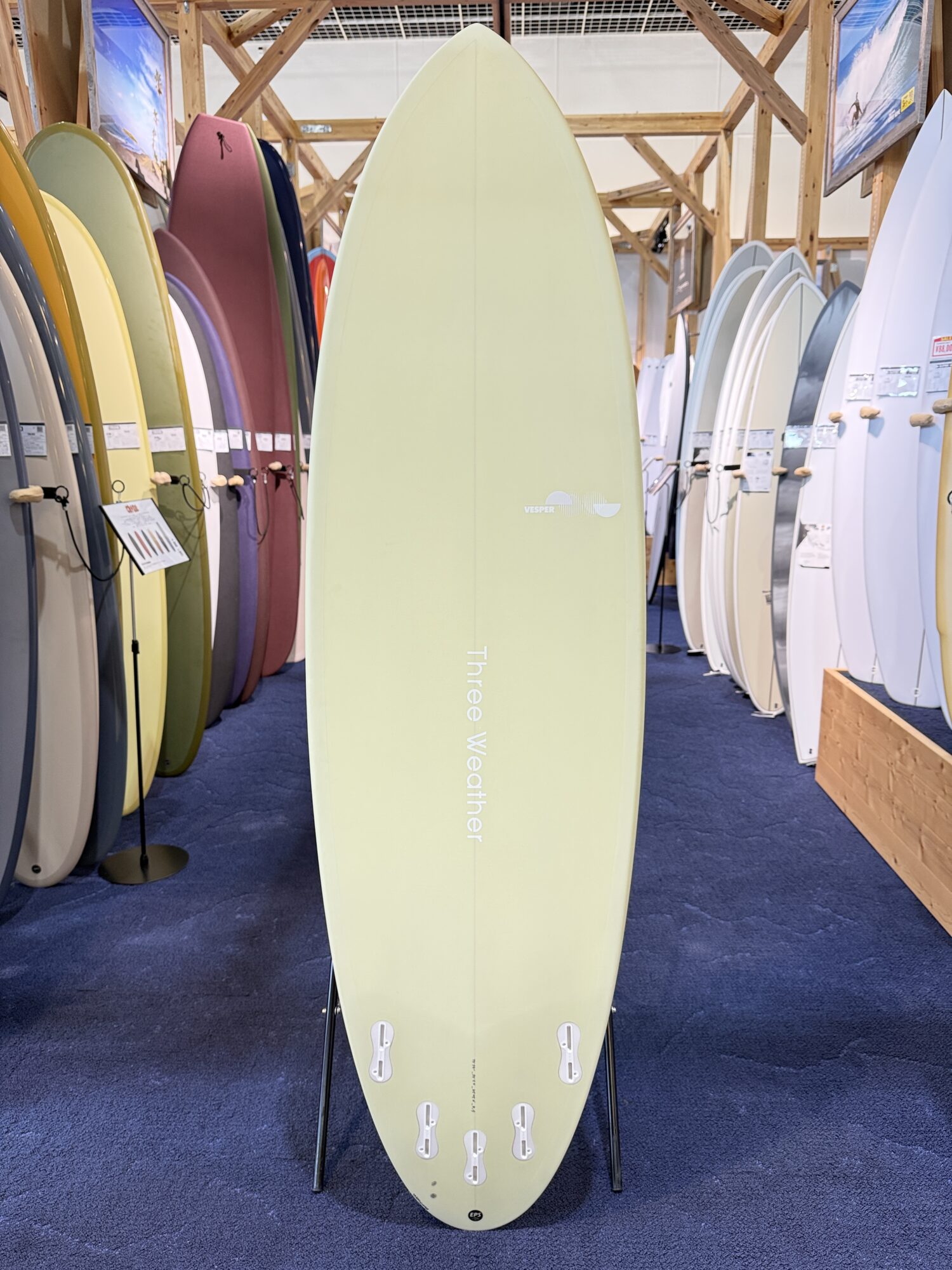 IMG_4024 - SHOP NEWS | ムラサキスポーツオンラインストア 通販 Pale yellow surfboard on display stand in a showroom, surrounded by tall colorful boards and wooden racks.