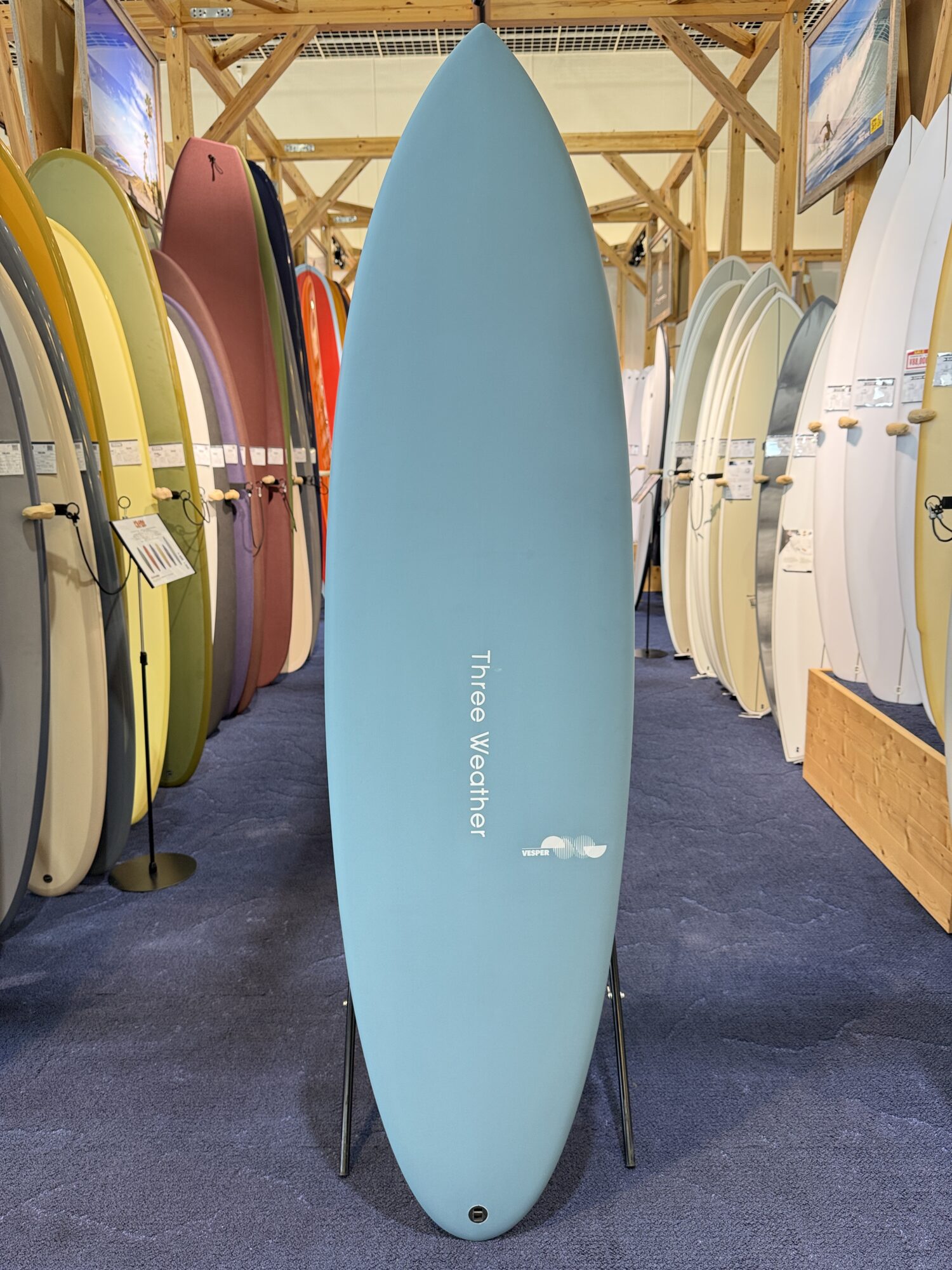 IMG_4025 - SHOP NEWS | ムラサキスポーツオンラインストア 通販 Blue surfboard standing upright on a stand in a showroom, flanked by rows of colorful boards on wooden racks.