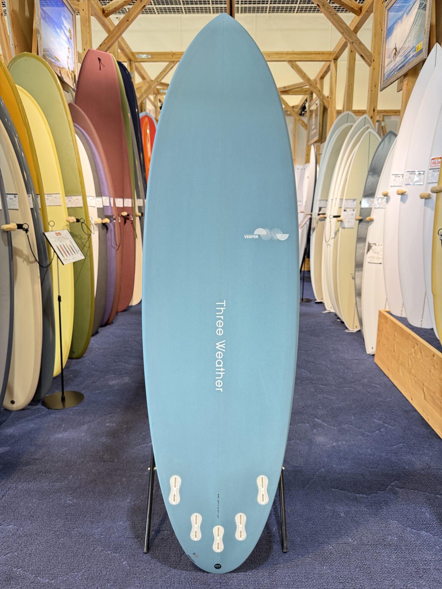 IMG_4026 - SHOP NEWS | ムラサキスポーツオンラインストア 通販 Blue longboard surfboard on a stand in a surf shop, surrounded by colorful boards on racks in a showroom.