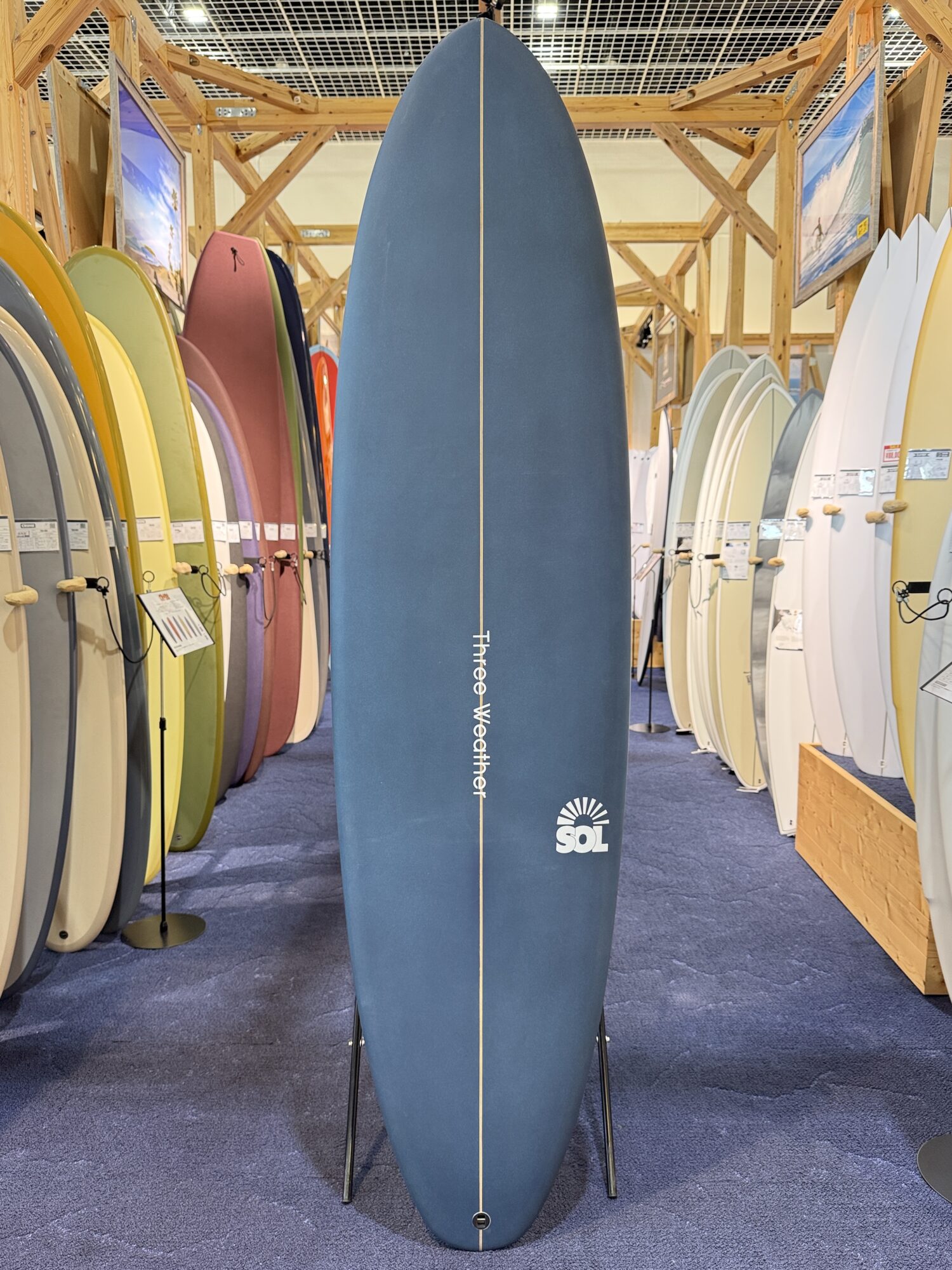 IMG_4027 - SHOP NEWS | ムラサキスポーツオンラインストア 通販 Tall blue surfboard on a display stand in a surf shop, surrounded by rows of colorful boards on both sides.