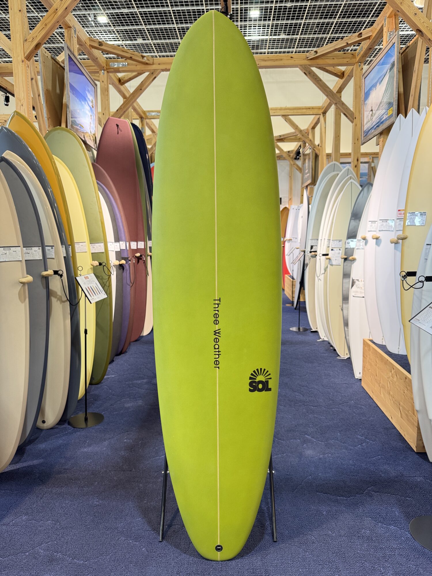 IMG_4029 - SHOP NEWS | ムラサキスポーツオンラインストア 通販 Indoor surfboard showroom with a lime-green board centered among colorful boards on racks; SOL logo near bottom.