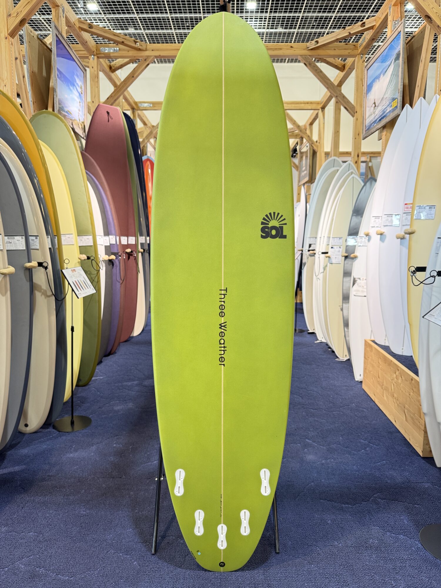 IMG_4030 - SHOP NEWS | ムラサキスポーツオンラインストア 通販 Bright lime-green surfboard centered in a shop with rows of colorful boards on both sides, wall posters in background.