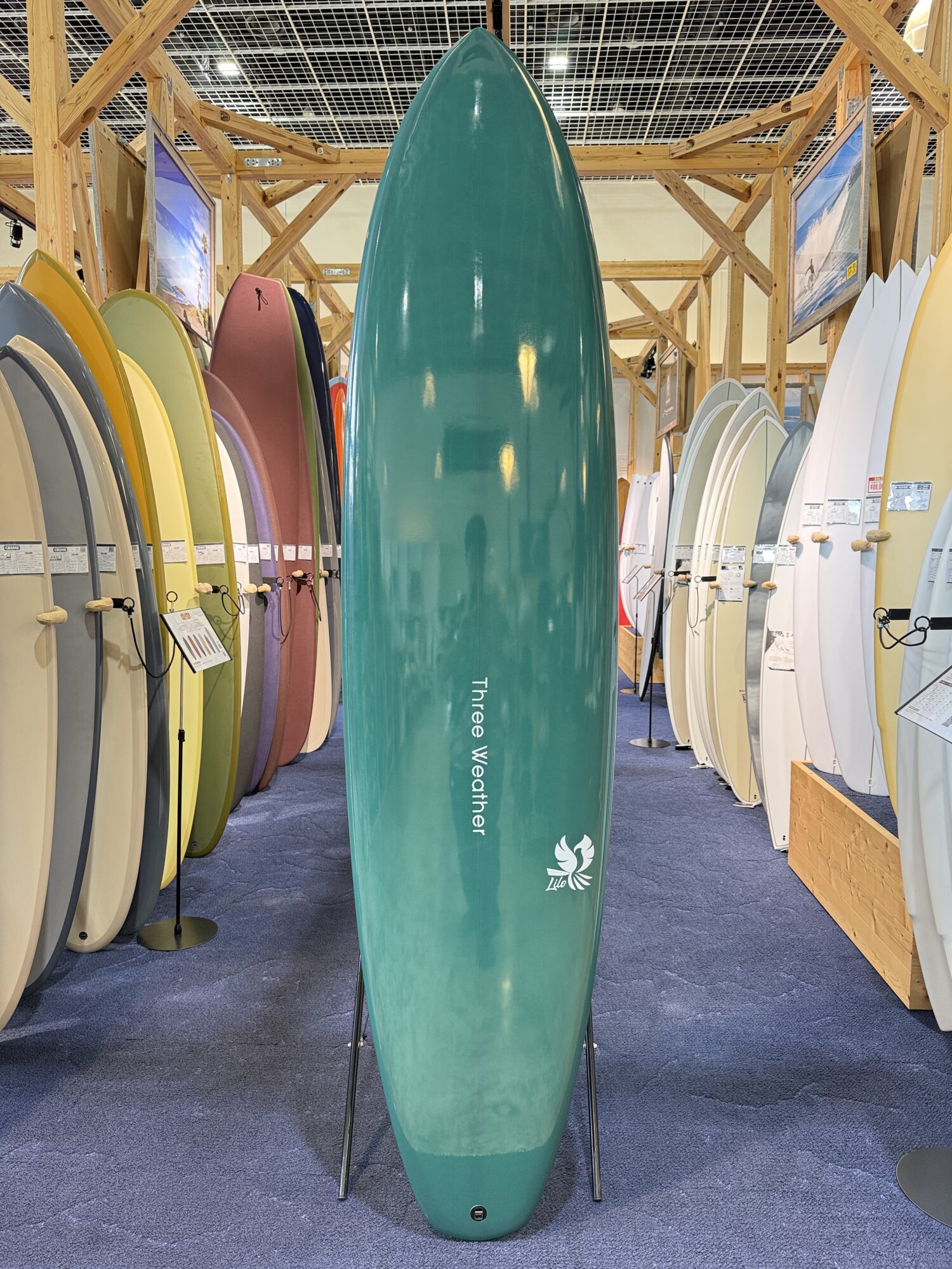 IMG_4031 - SHOP NEWS | ムラサキスポーツオンラインストア 通販 Teal longboard standing upright on a stand in a surf shop, surrounded by colorful surfboards on racks inside a wooden-framed showroom.