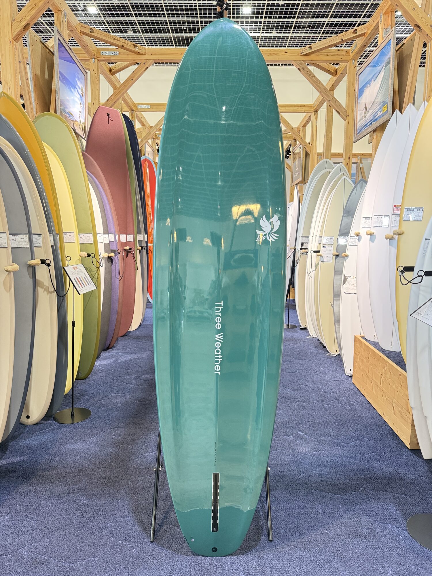 IMG_4032 - SHOP NEWS | ムラサキスポーツオンラインストア 通販 Teal surfboard on display stand in a showroom, flanked by rows of colorful boards on wooden racks.
