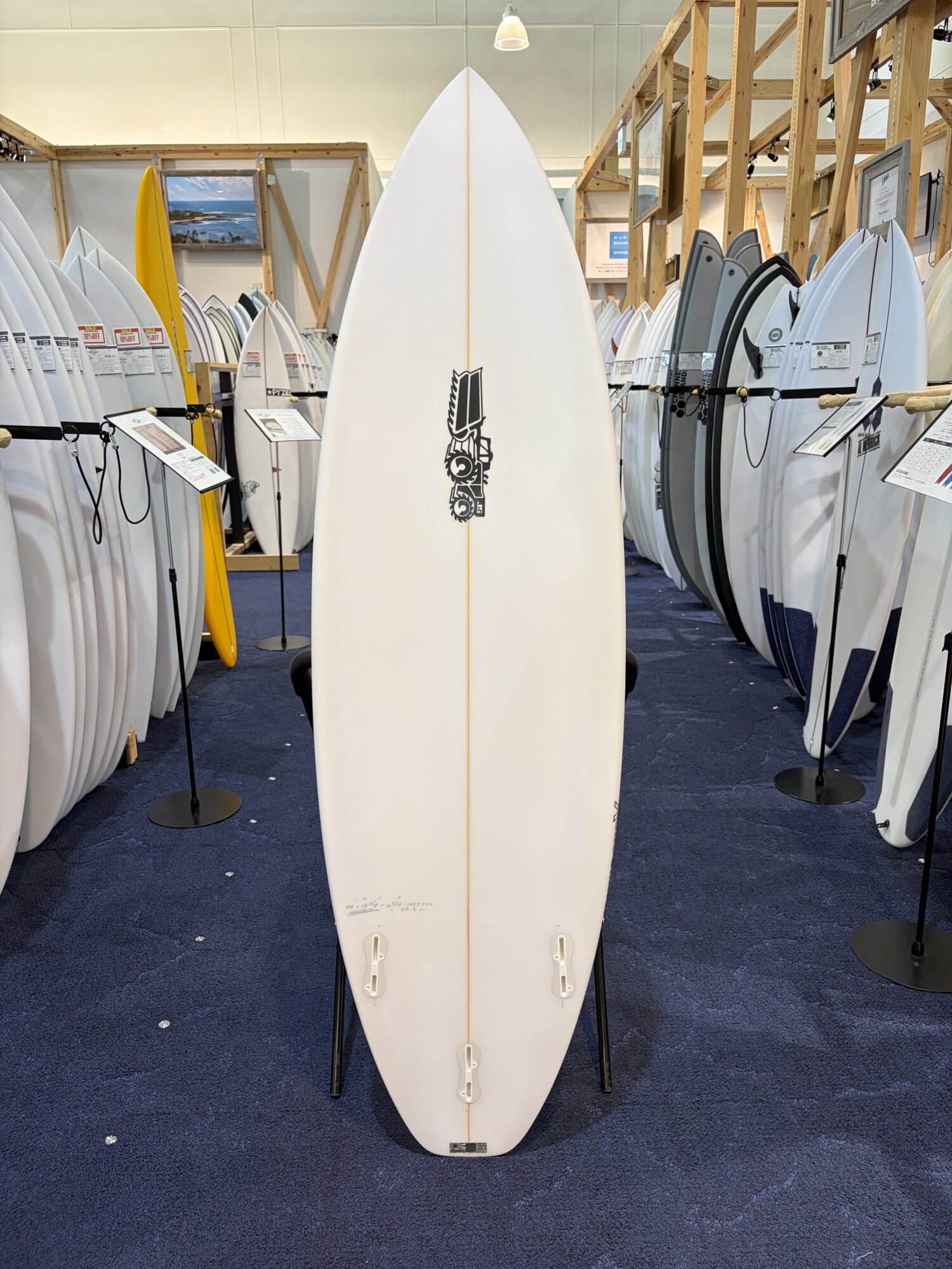 Tall white surfboard on a stand in a showroom, surrounded by racks of other surfboards on both sides.