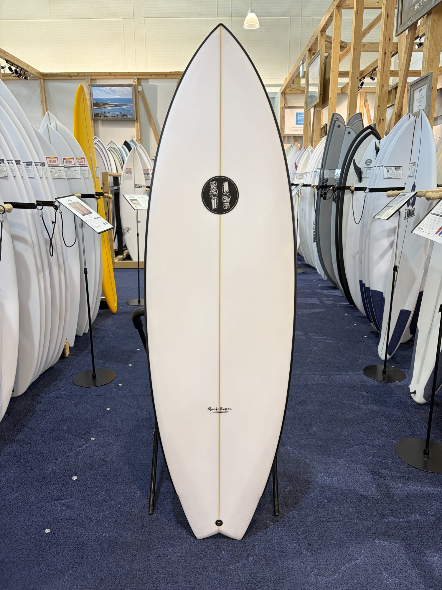 White surfboard on a display stand in a shop, with rows of surfboards lined up on both sides in the background.