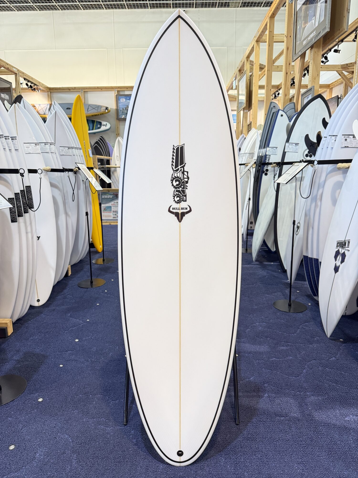 Tall white surfboard with a black edge standing on a stand in a surf shop, surrounded by other boards; central logo resembles a stylized figure with 'BULL RUN' at the bottom.