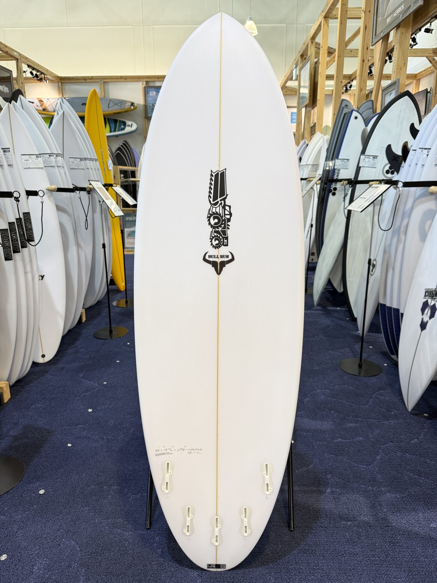 White surfboard on a stand in a showroom, centered with a Bull Run logo and a yellow stringer visible down its length.