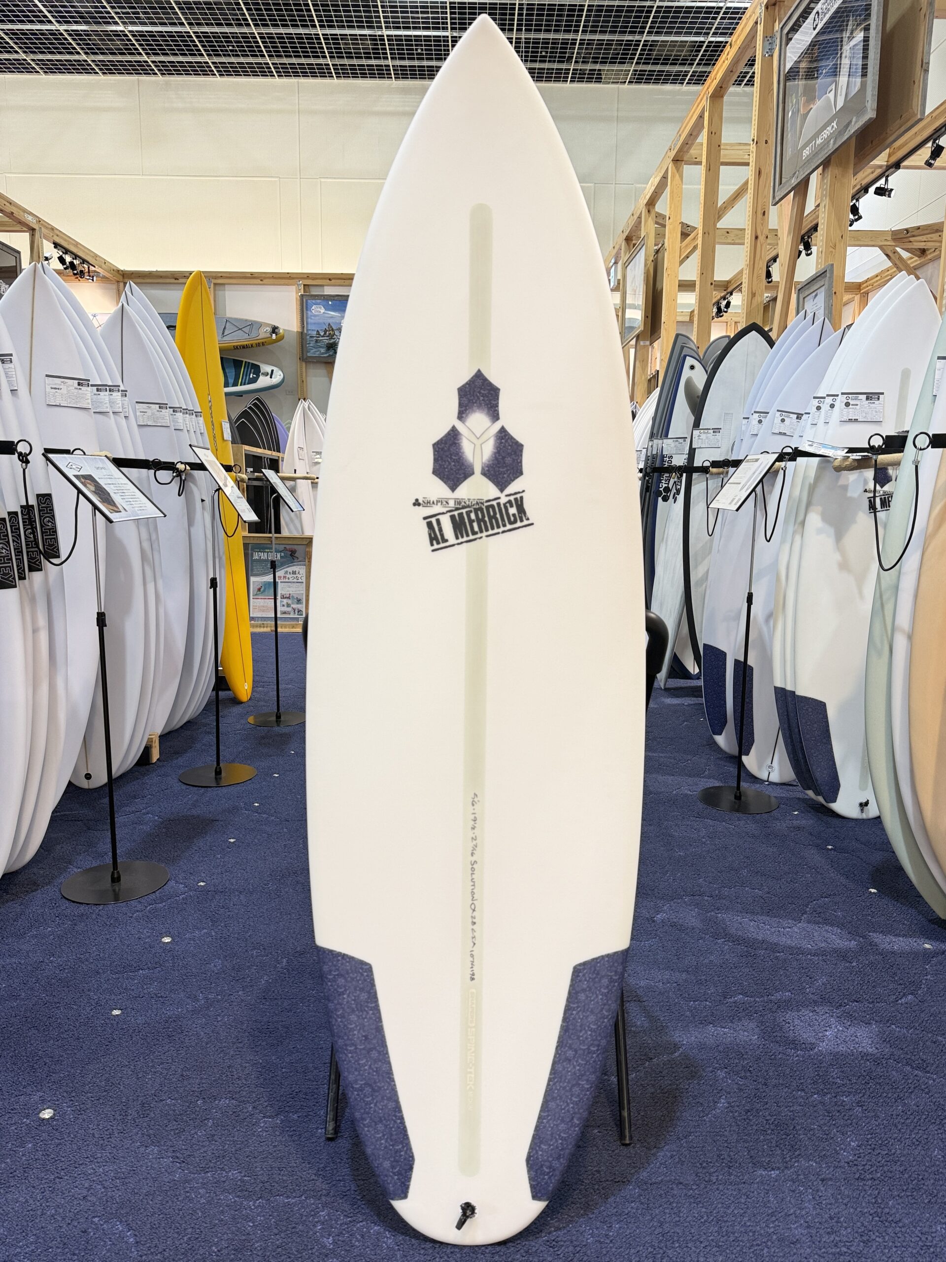 IMG_7480 - SHOP NEWS | ムラサキスポーツオンラインストア 通販 Central white surfboard with the Al Merrick logo stands upright in a showroom, surrounded by numerous other boards on display in wooden racks.