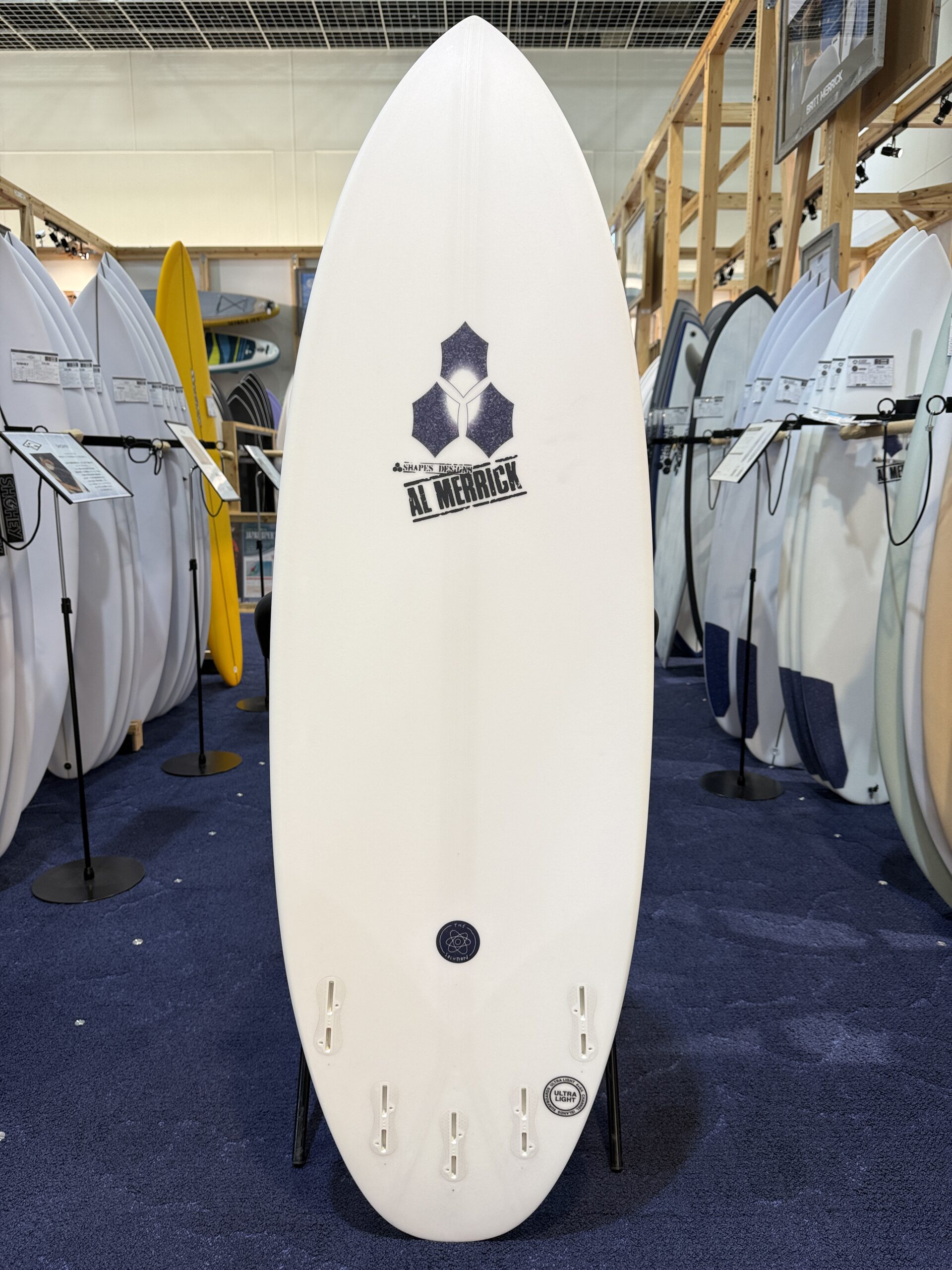 IMG_7481 - SHOP NEWS | ムラサキスポーツオンラインストア 通販 White surfboard standing upright in a showroom, surrounded by other boards; centered logo reads 'AL MERRICK' with a blue geometric emblem.