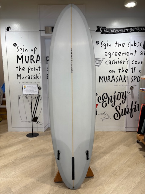 White surfboard standing upright on a wooden rack indoors, with a faint orange center stringer and two fin boxes visible at the bottom.